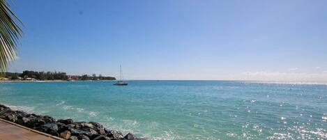A coastal view with a rocky shoreline, clear blue water, and a sailboat in the distance.