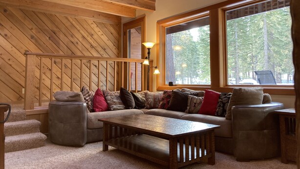 Ein gemütliches Wohnzimmer mit Holzbalkendecke, einem großen Fenster mit Blick auf einen Wald, einer Couch mit gemusterten Kissen und einem Holztisch.