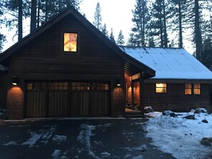 Una cabaña de madera con garaje, una ventana iluminada y techo cubierto de nieve.