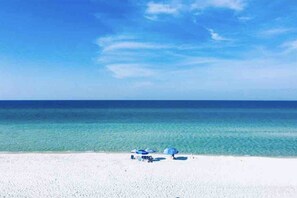 A beach with white sand and a clear blue sky. There are two blue umbrellas and a few beach chairs. - A+ Location across from beach. Free bikes & beach equipment! (Santa Rosa Beach)