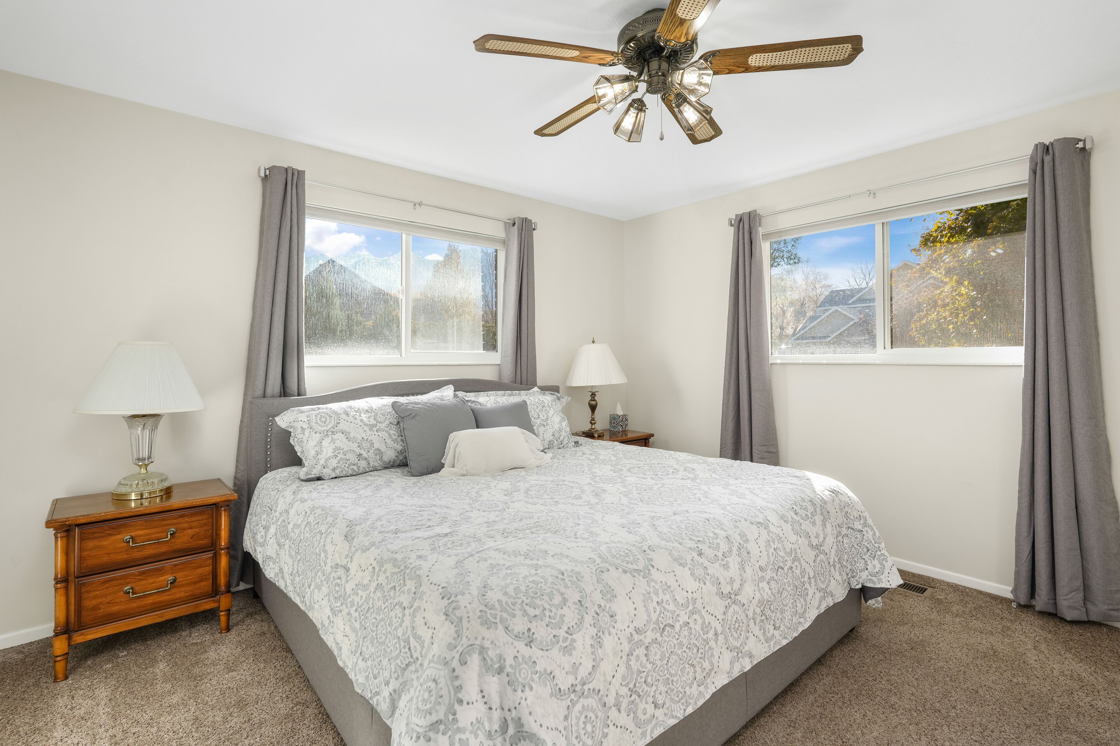 A bedroom with a bed, nightstands, lamps, a ceiling fan, and two windows with curtains.