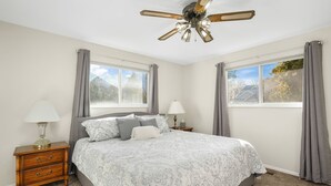 Un dormitorio con cama, mesitas de noche, lámparas, ventilador de techo y dos ventanas con cortinas.