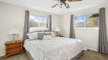 A bedroom with a bed, nightstands, lamps, a ceiling fan, and two windows with curtains.