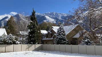 A snow-covered landscape with a house featuring a stone facade and a traditional design, surrounded by snow-laden trees and mountains in the background.