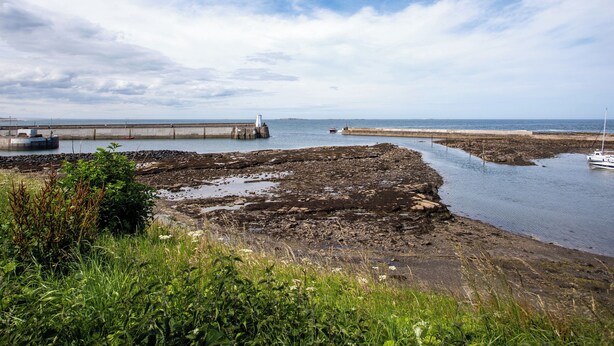 A coastal landscape with a rocky shoreline, a pier extending into the water, and a boat anchored near the shore. - Harbour Lights House (Seahouses)