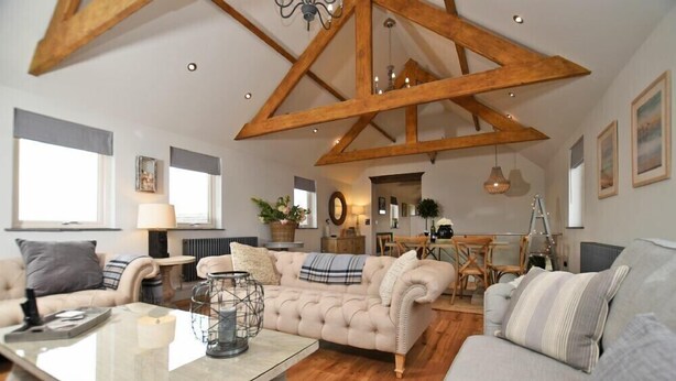 A spacious living room with a high ceiling, exposed wooden beams, a central seating area with a sofa and armchairs, a coffee table, and a dining area in the background. - The Hemmel (Chathill)