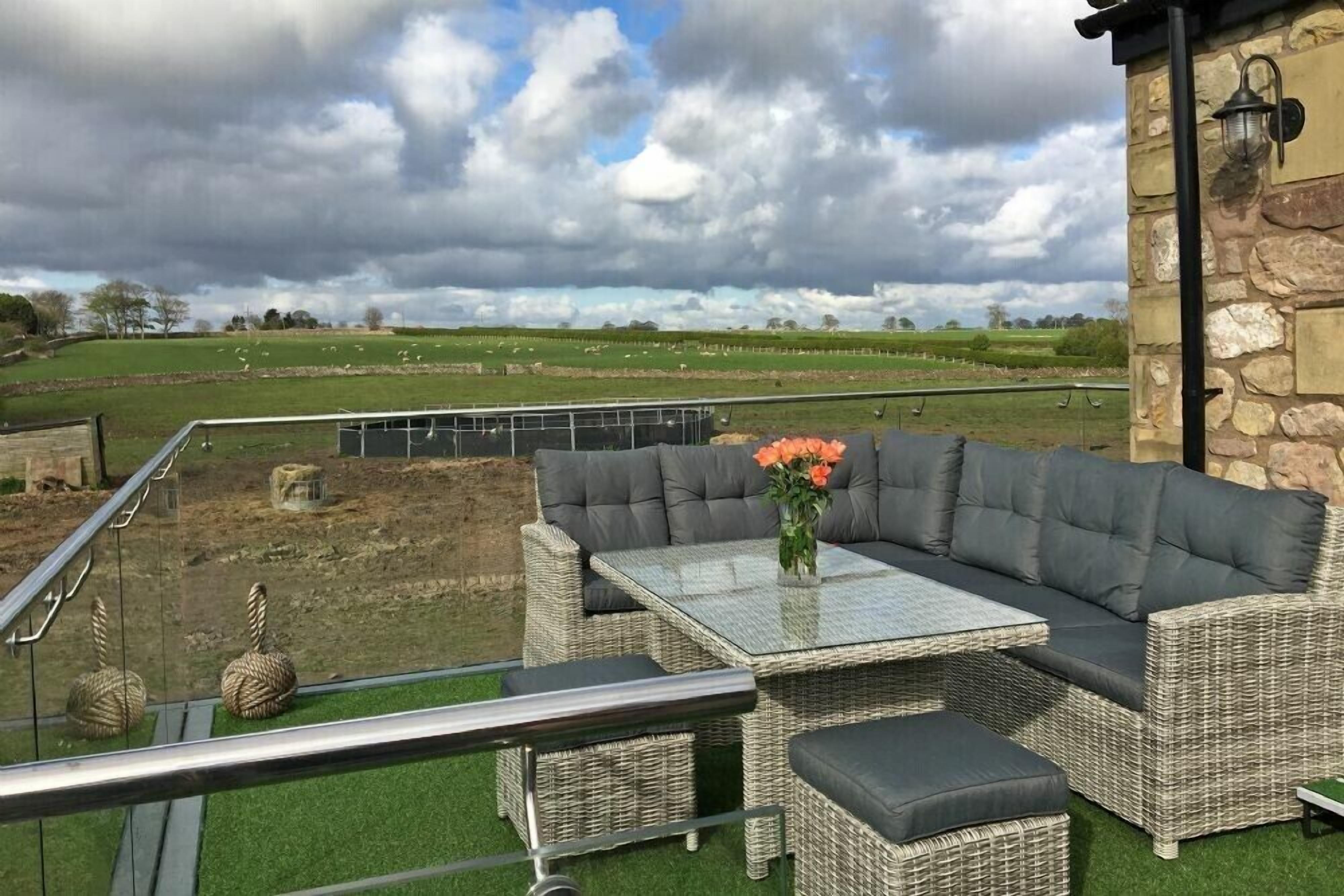 A balcony with wicker furniture, a glass table, and a view of a green field with sheep.