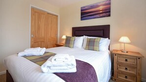 A neatly made bed with white linens and a purple throw blanket, a wooden door, a nightstand with a lamp, and a painting of a sunset on the wall.