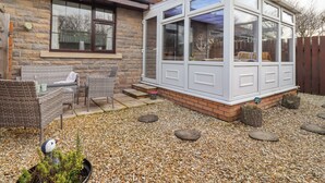 A house with a stone exterior, a covered patio with wicker furniture, and a large windowed sunroom. - The Bolthole (Seahouses)