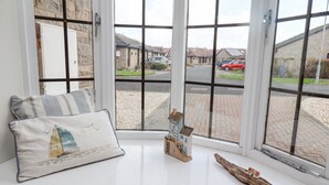 A room with a large window, a white bench with a cushion, and a small model house on the windowsill. - The Bolthole (Seahouses)