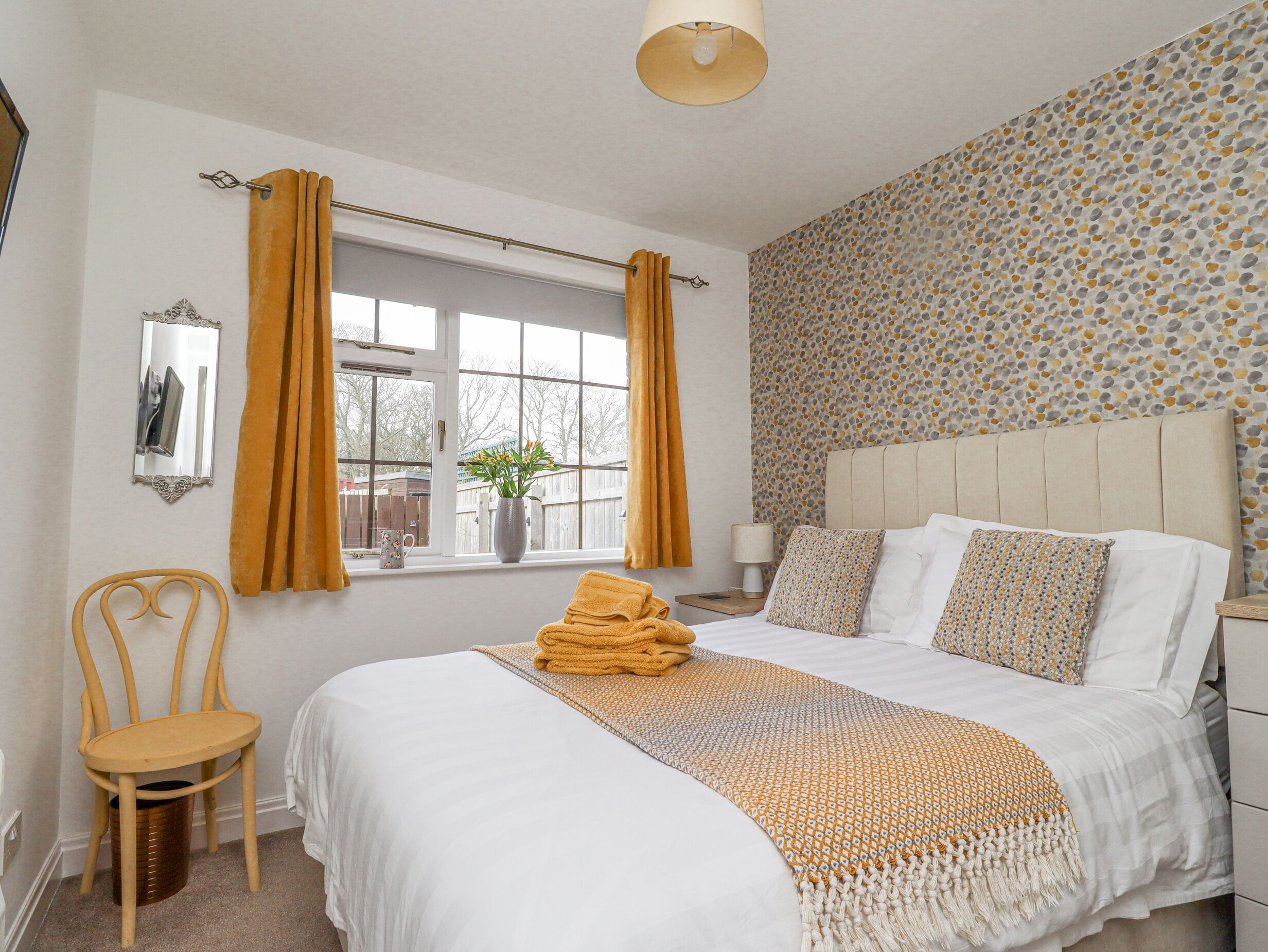 A bedroom with a bed, a chair, a window with curtains, a bedside table, and a wall with a patterned wallpaper.