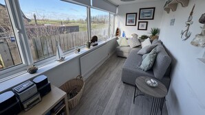A cozy living room with a grey sofa, a small round table, and a large window overlooking a backyard.