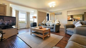 Een moderne woonkamer met een flatscreen televisie, een houten salontafel, grijze banken en uitzicht op de omgeving door grote ramen.