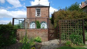 Una casa torre de ladrillo con una ventana pequeña y una puerta, rodeada por un jardín con valla de madera y una pérgola.