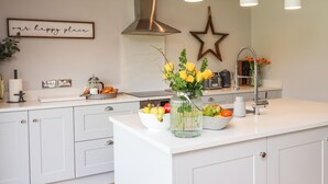 Una cocina moderna con armarios blancos, una isla central con fruta y flores, una campana extractora de acero inoxidable, luces colgantes y un adorno de estrella en la pared.
