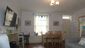 Sala de estar com lareira, área de jantar com cadeiras de madeira, sofá cinza e escada à direita.