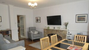 Uma sala de estar com lareira, televisão fixada na parede e área de jantar com cadeiras de madeira e mesa de vidro.