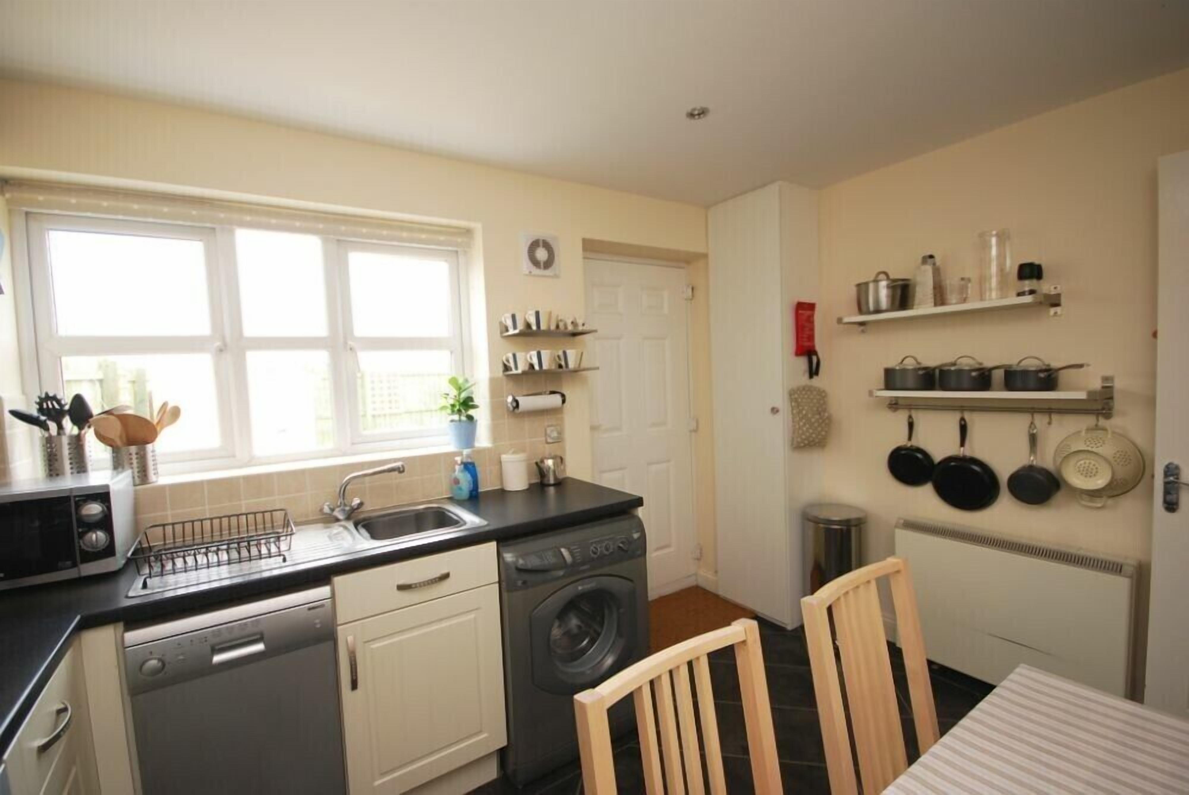 A compact kitchen with a dishwasher, washing machine, and a sink. There are shelves above the sink and a chair at the table.