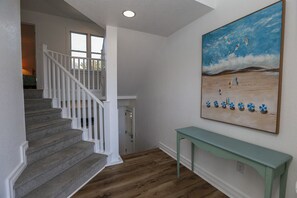 A staircase with white railings leading to a landing, a painting of surfers on a beach, and a teal console table.