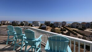 A deck with four turquoise Adirondack chairs overlooking a coastal residential area with houses and the ocean.
