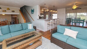 A spacious living area with two teal sofas, a wicker coffee table, and a kitchen area with white cabinets and a bar. - By Jove - Semi-Oceanfront 5 BR Beach Cottage w/Private Pool & Dogs Allowed (Southern Shores)