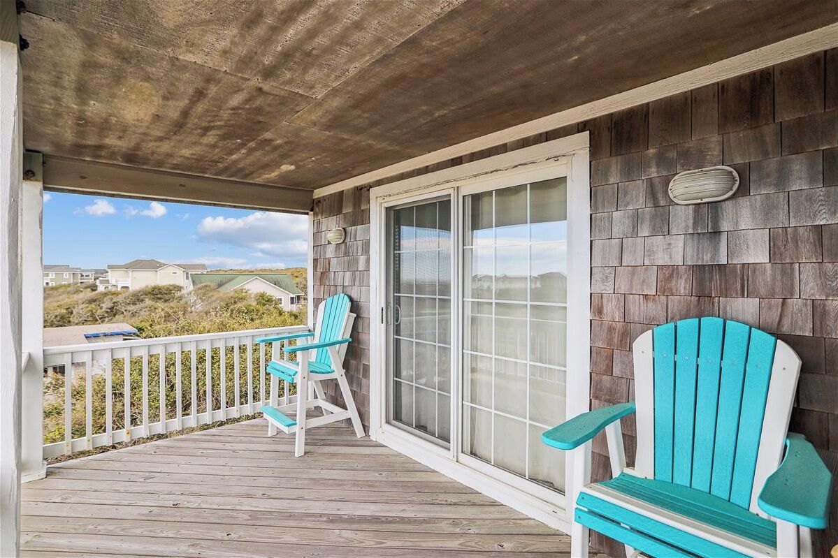 A covered porch with two turquoise Adirondack chairs, a white railing, and a view of the beach and houses in the distance.