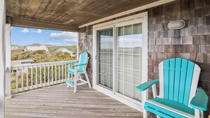 A covered porch with two turquoise Adirondack chairs, a white railing, and a view of the beach and houses in the distance.