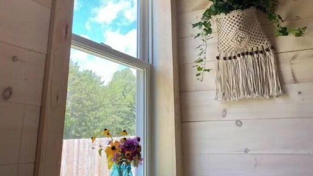 A room with wooden walls, a window with a view of the sky and greenery, a hanging macrame wall decoration, and a vase with flowers on the windowsill.