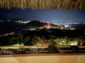 ’s Nachtzicht vanaf een balkon met rieten dak, met uitzicht op een stadsbeeld met verlichte gebouwen en een heuvel in de verte.