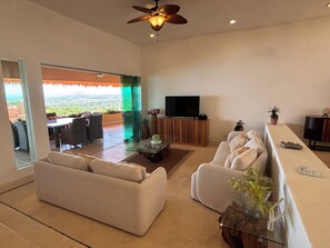 A spacious living area with a sofa, a coffee table, and a television. There is a balcony with a view of the landscape.