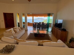 A living room with a sofa, a coffee table, and a television on a wooden cabinet. There is a view of the outdoors through large windows. - Great luxurious penthouse with private roof top pool lounge and spectacular view (Ixtapa Zihuatanejo)
