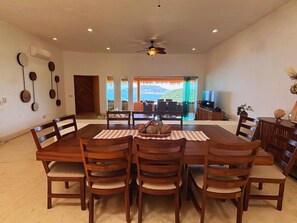 Un comedor con una gran mesa de madera, varias sillas y vista al océano a través de un ventanal.