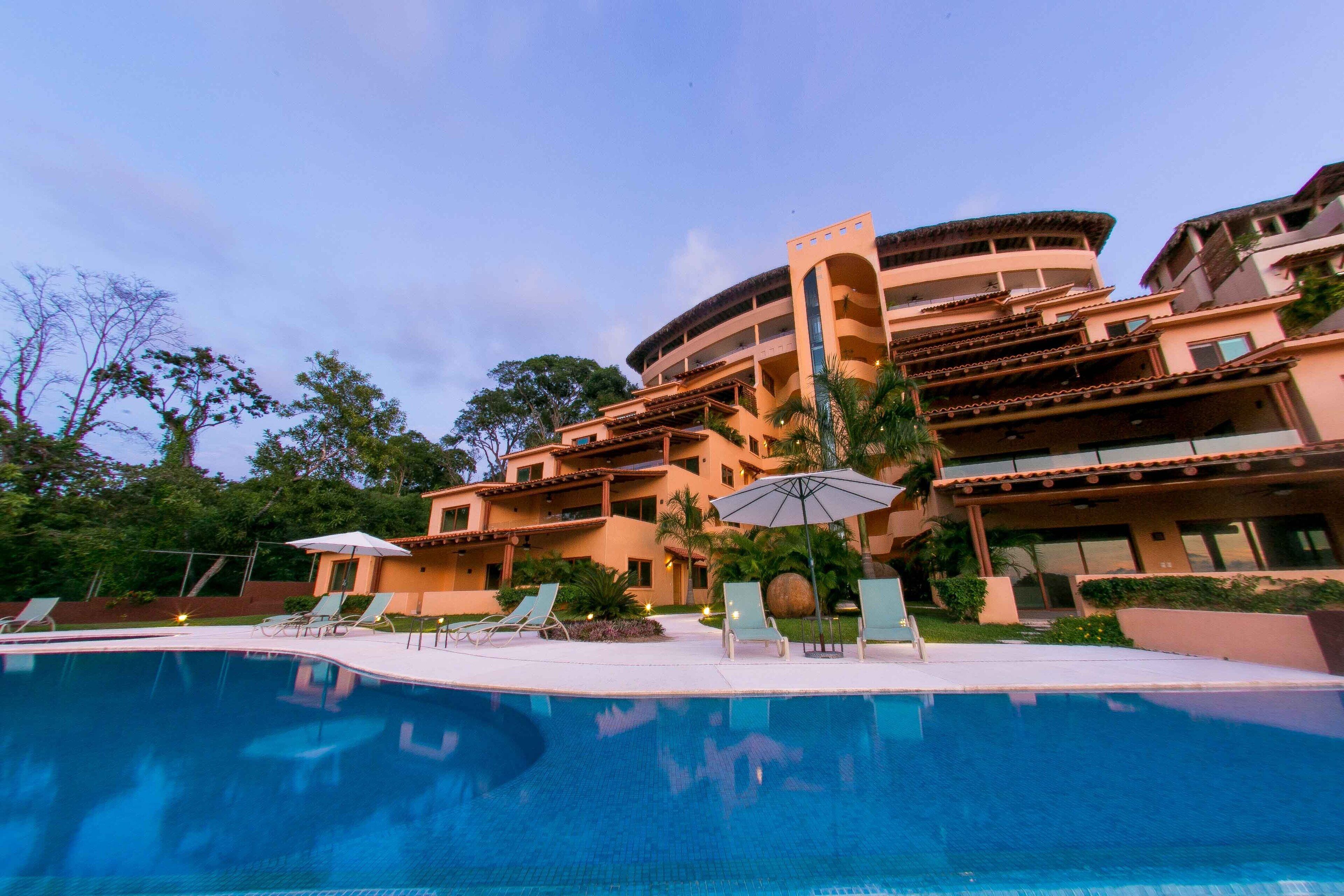 A hotel with a swimming pool, lounge chairs, and umbrellas.