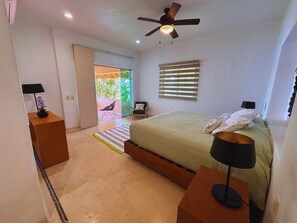Un dormitorio con una cama, dos mesitas de noche, un ventilador de techo, una ventana con persianas y una puerta que da a un balcón.