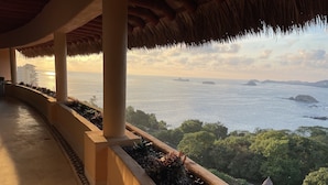 Un balcon avec vue sur la mer, des palmiers et une structure surmontée d’un toit de chaume.