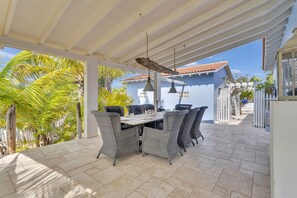 A covered patio with a dining table and chairs, surrounded by palm trees and a building with a tiled roof.