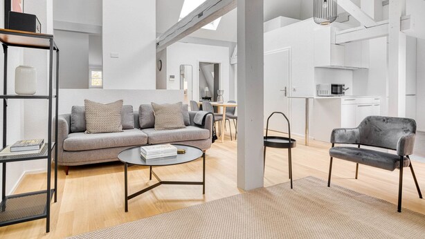 A spacious living area with a grey sofa, a round coffee table, and a black armchair. There is a bookshelf with books and a vase, and a kitchen area in the background. - Chic Vorstädte 2.5 Room w/ W/D & Storage, 2 min to Market, by Blueground (Basel)