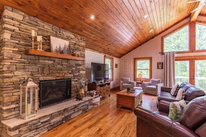 A cozy living room with a stone fireplace, wooden ceiling, and large windows.