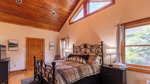 Un dormitorio con techo de madera, una cama con estructura metálica, una mesita de noche con una lámpara y una ventana con vista a árboles.