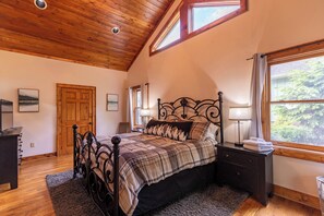 A bedroom with a wooden ceiling, a bed with a metal frame, a nightstand with a lamp, and a window with a view of trees.