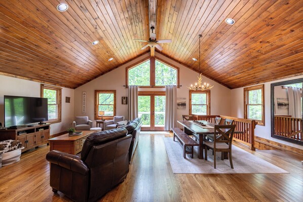 A spacious living area with a high ceiling, wooden beams, a large television, a leather sofa, a dining table with chairs, and a chandelier.