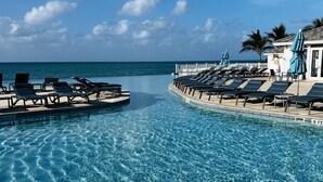 Ein klares, blaues Schwimmbecken mit Liegestühlen und Sonnenschirmen, umgeben von einem weißen Zaun und Palmen, mit Blick auf den Ozean.