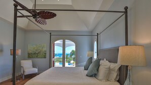 Un lit à baldaquin avec vue sur la mer, visible à travers une porte vitrée, un tableau accroché au mur et une lampe sur la table de chevet.