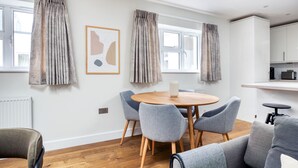 A modern living room with a round wooden table, grey upholstered chairs, a grey sofa, and a kitchen area with white cabinets and a black countertop. - Stylish Marylebone 2BR nr Underground, by Blueground (London)