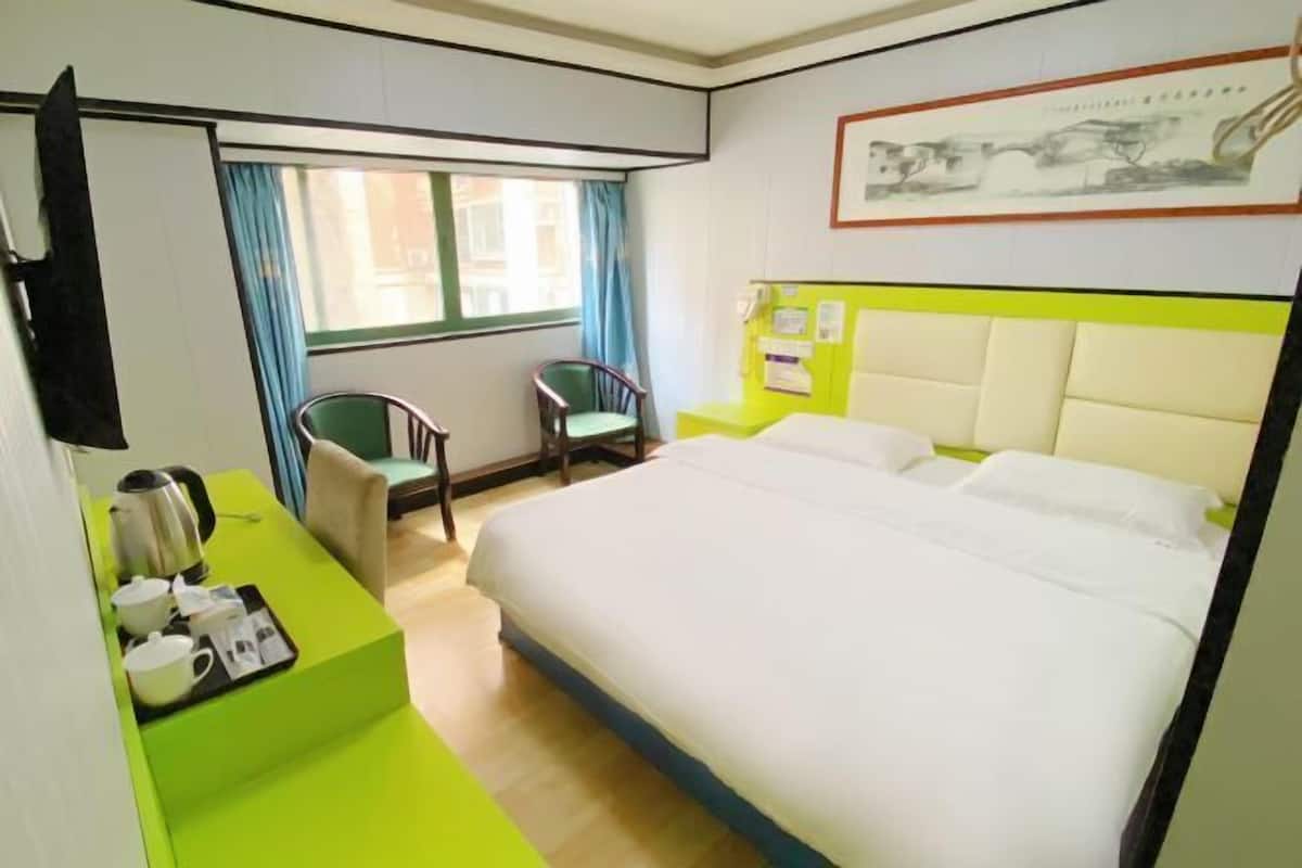 A hotel room with a bed, a desk with a kettle, a chair, and a window with curtains.