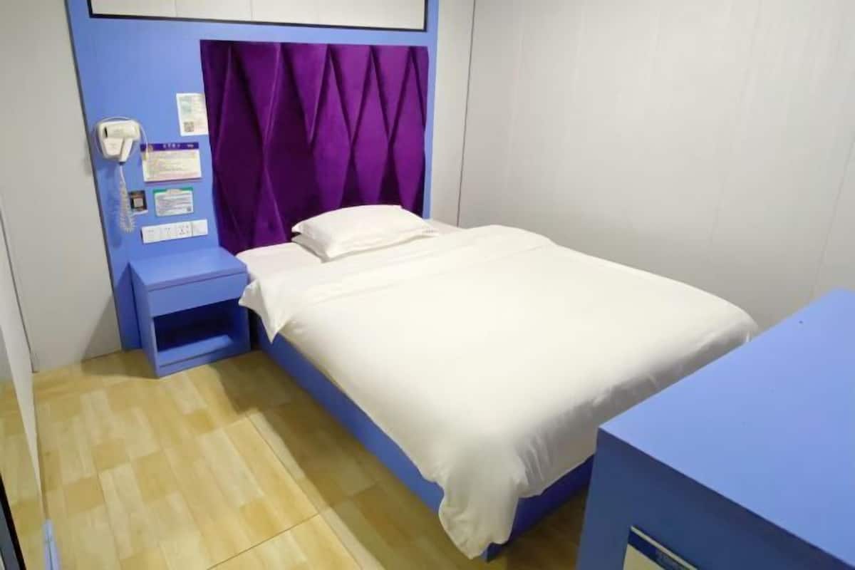 A single bed with a white mattress and a purple headboard, a blue nightstand, and a wall-mounted phone.