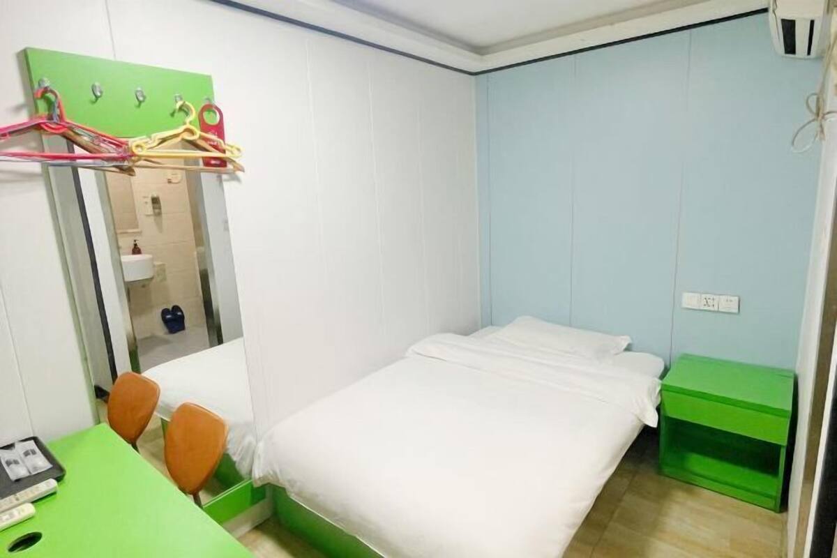 A small, neatly organized room with a bed, a desk, and a chair. There is a mirror on the wall, and a green storage unit is visible.