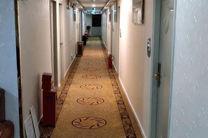 Een hotelgang met een tapijtvloer, deuren aan weerszijden en een brandblusser aan de rechtermuur.