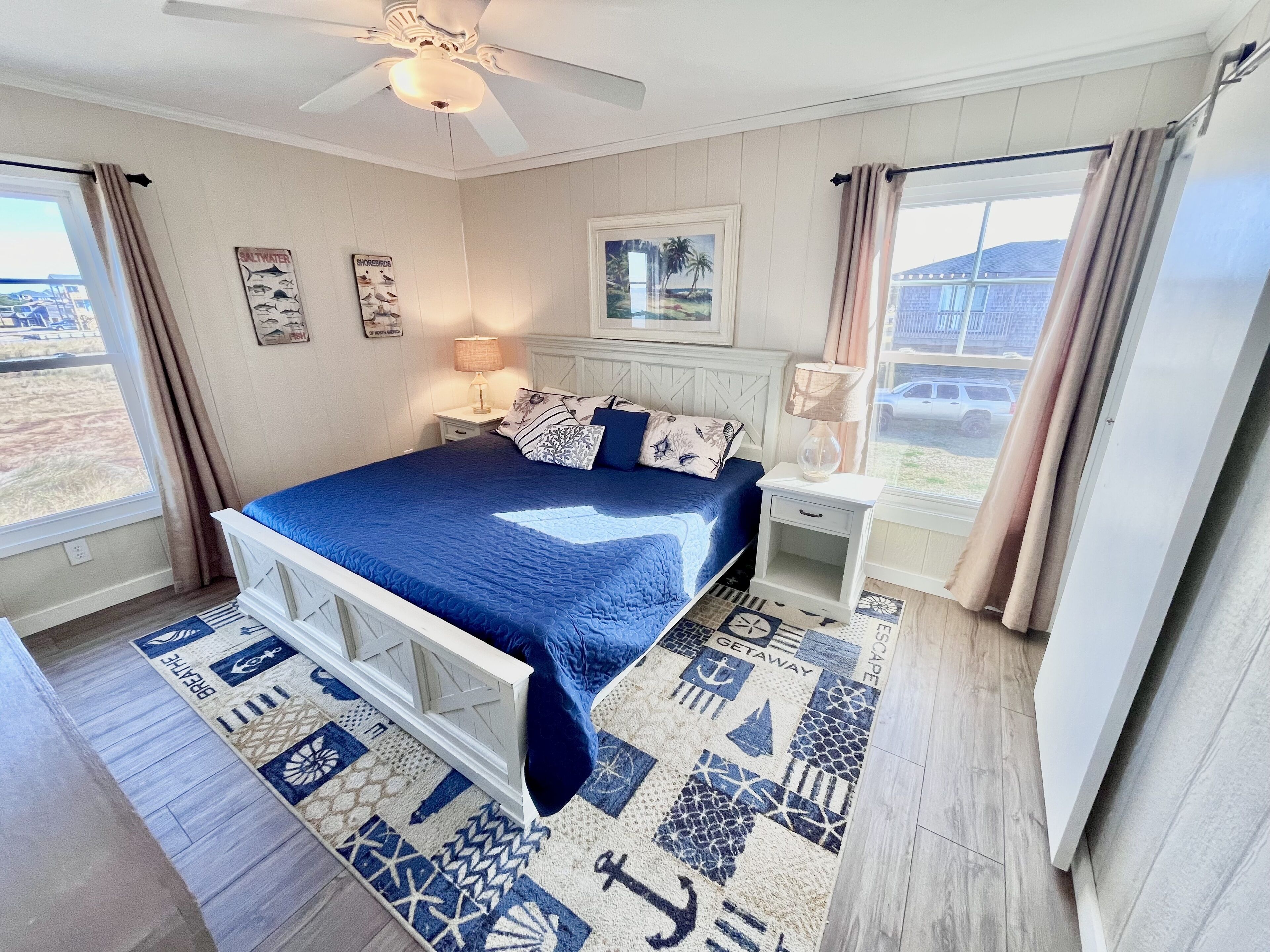 A bedroom with a bed, a nightstand, a ceiling fan, a window with curtains, and a view of the outside.