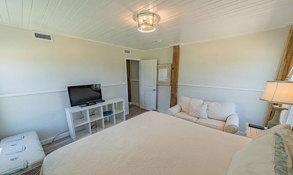 A bedroom with a large bed, a TV on a white stand, a white sofa, and a lamp.